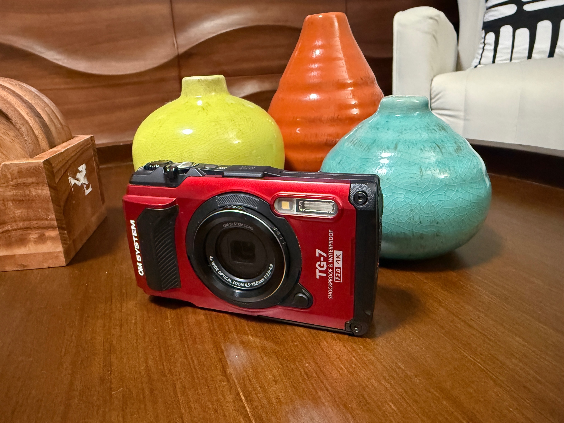 OM SYSTEM TG-7 in red — waterproof, shockproof adventure camera resting on a wooden table beside yellow, orange, and turquoise ceramic vases
